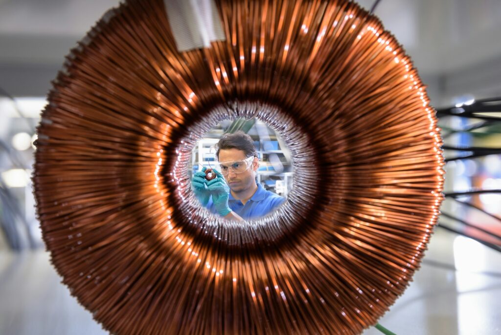 Worker inspecting electromagnetic coil seen through large coil in electromagnetics factory